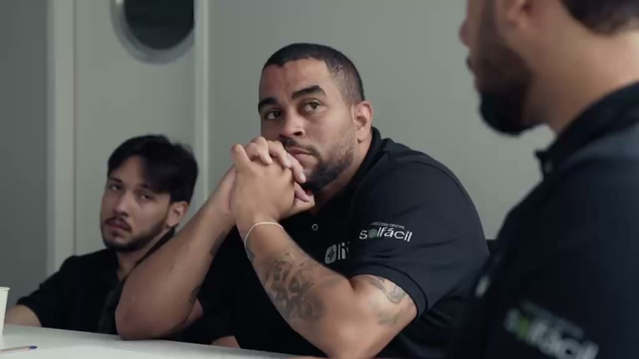 Reunião numa sala de escritório: ao centro, um homem de camisa polo preta com o logo da Solfácil escuta com as mãos cruzadas, com dois integrantes da equipe ao lado.